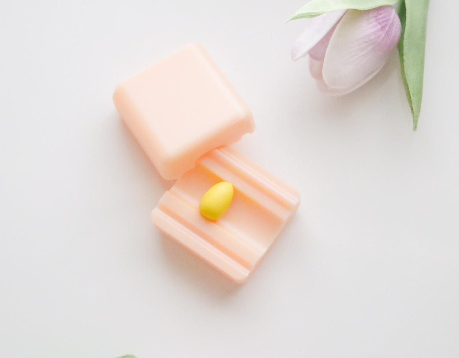 A small yellow pill sits in an open pale pink case on a white surface, with a light purple tulip flower nearby.
