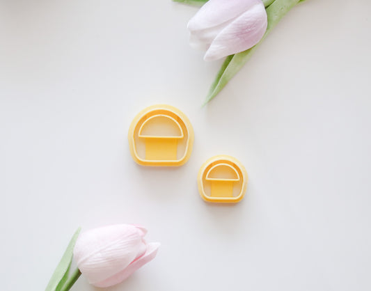 Two pastel pink tulips with green stems frame two yellow, mushroom-shaped Hello Cutters Basket Polymer Clay Cutter STL Set pieces in different sizes on a white background—ideal for a unique digital download.