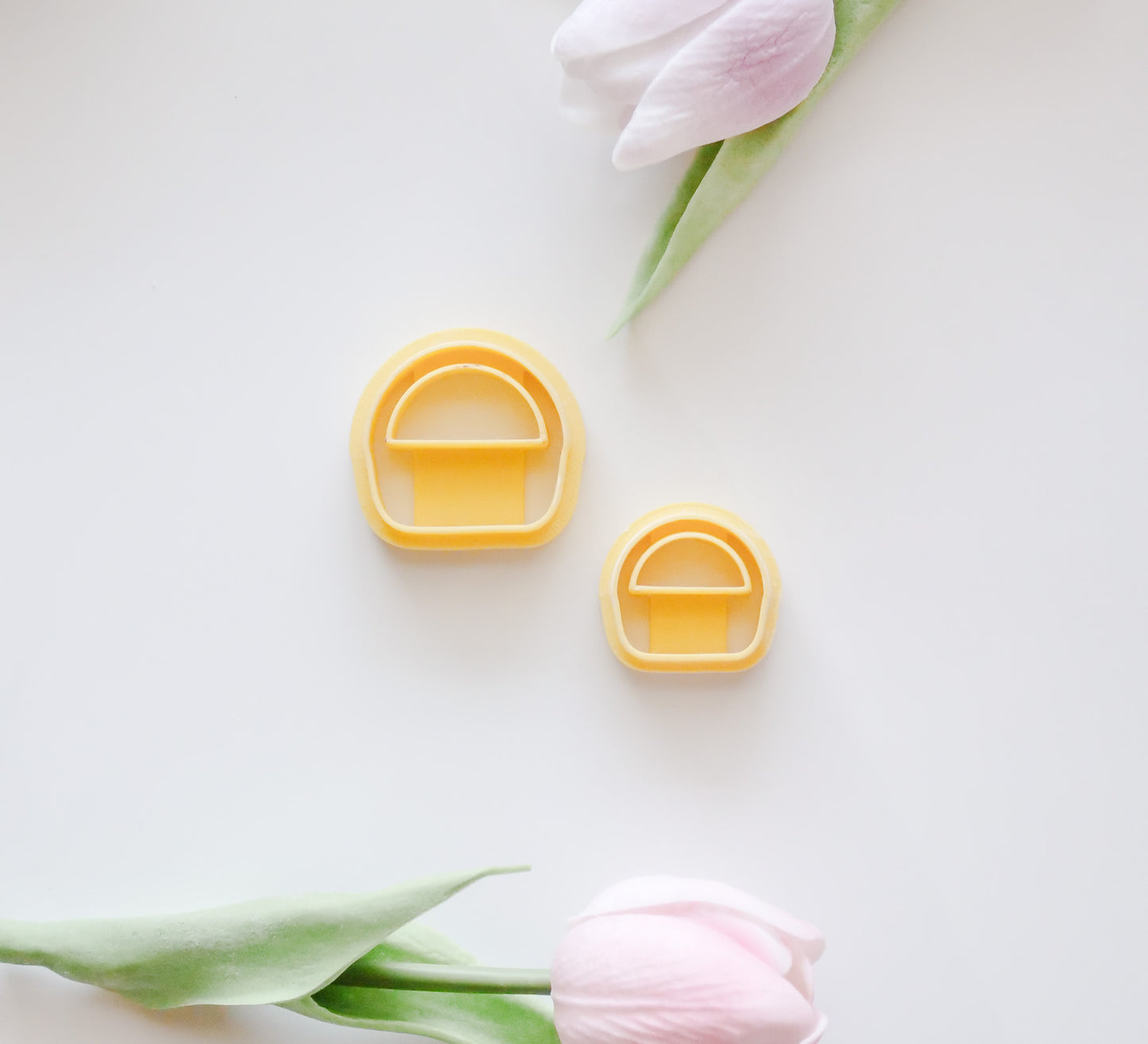 The Hello Cutters Basket Polymer Clay Cutter STL Set includes two yellow, mushroom-shaped cutters (large and small), displayed on a white surface with pink tulips—ideal for digital download and creative projects.