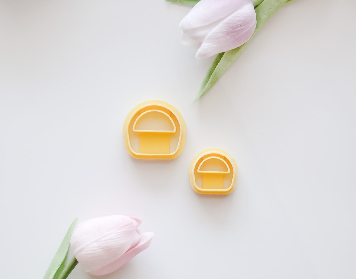 Two pastel pink tulips with green stems frame two yellow, mushroom-shaped Hello Cutters Basket Polymer Clay Cutter STL Set pieces in different sizes on a white background—ideal for a unique digital download.