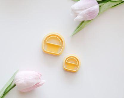 Two yellow, dome-shaped Basket Polymer Clay Cutter STL Set cutters with half-circle cutouts by Hello Cutters are centered on a white surface framed by pink tulips—perfect for your next digital download project.