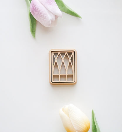 The Hello Cutters Window Polymer Clay Cutter Set, featuring a rectangular, beige cutter with an arched window pattern, is displayed on a white surface alongside a pink tulip above and a yellow tulip below.